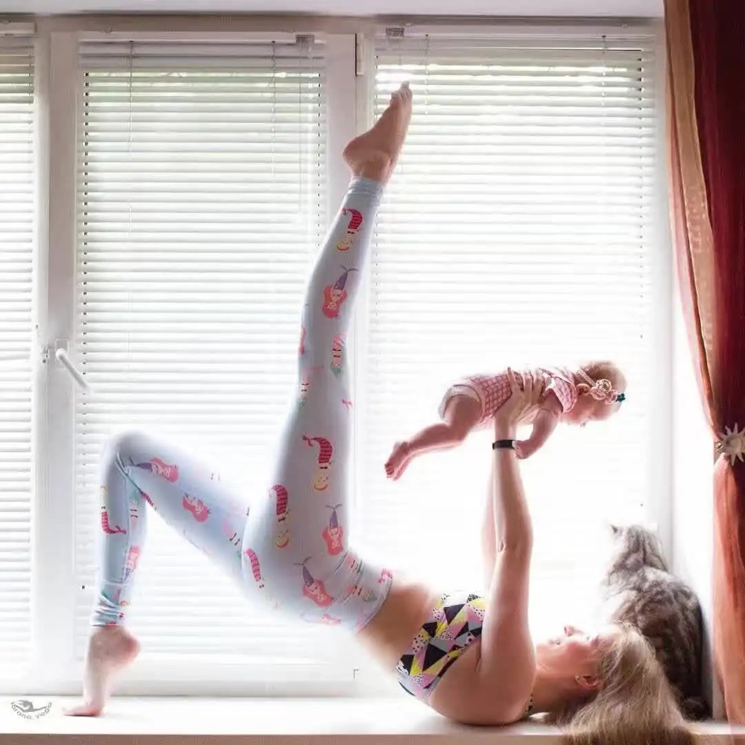 Woman practicing yoga in bright patterned yoga pants, holding a baby in a playful pose.